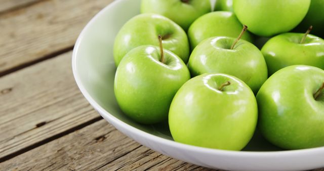 Displays fresh, green apples in a white bowl on rustic wooden table, presenting an image perfect for marketing health foods, promoting an organic lifestyle, and suggesting natural snacks. Use in blogs, websites, or social media posts promoting healthy eating.