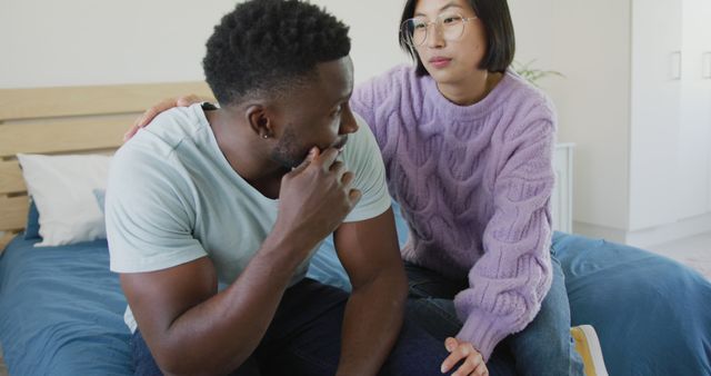 Shows an interracial couple sitting closely and sharing a quiet, serious moment, with one person offering comfort by placing a hand on the other's shoulder. Ideal for use in articles or campaigns tackling themes related to relationships, emotional support, empathy, mental health, interracial harmony, and intimacy. May also be useful in counseling resources that address challenges faced by couples.