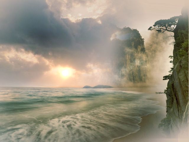 Capturing a dramatic coastal landscape during sunset with misty conditions and towering rocky cliffs. The scene showcases the beauty and tranquility of nature with calm ocean waves gently reaching the shore. This image is perfect for serene and peaceful backdrops, ideal for travel brochures, meditation apps, and nature-inspired designs.