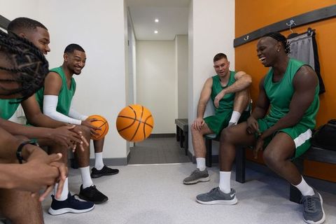 Diverse male basketball team laughing and tossing ball in green jerseys on locker bench
