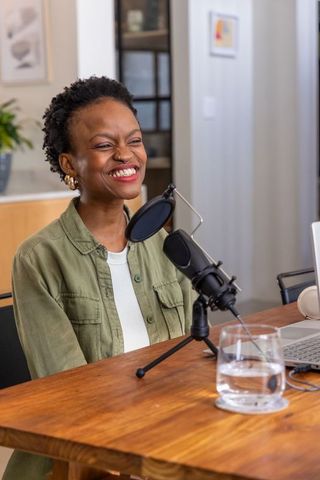 African american woman smiling while recording podcast in home studio with condenser microphone