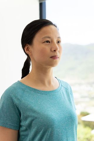 Serene woman standing by sunlit window wearing teal shirt and braid, contemplative portrait