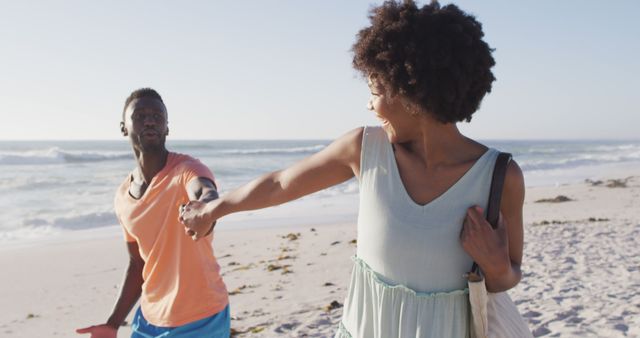 Smiling african american couple holding hands and walking on sunny beach. healthy, active family beach holiday.