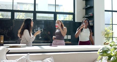 Three women socializing in bright modern kitchen holding drinks and checking tablet