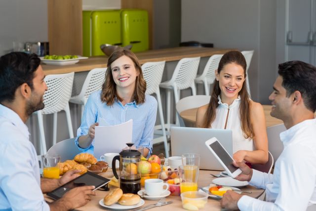 Business colleagues are having a breakfast meeting in an office cafeteria. They are discussing work while using digital tablets and laptops. The table is set with coffee, croissants, and fruits, creating a casual yet professional atmosphere. This image can be used for promoting corporate culture, teamwork, and modern work environments.