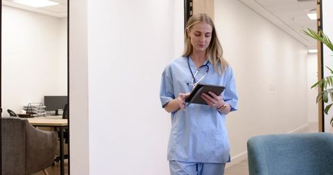 Female nurse reviewing tablet in modern clinic corridor with stethoscope