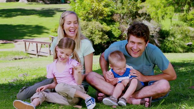 Family sitting together on grass in a park, parents playfully tickling their kids. Perfect for themes promoting family time, outdoor activities, bonding, and happiness. Suitable for advertisements, brochures, and posters highlighting family-centric products or services.