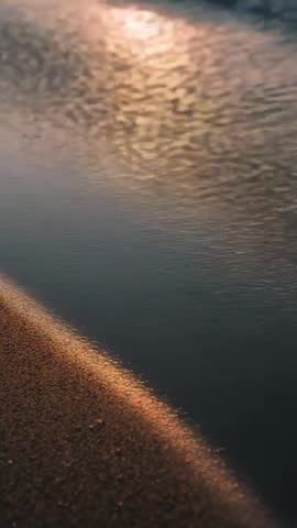 Vertical video showing golden hour waves lapping wet sand with sun glinting on ripples