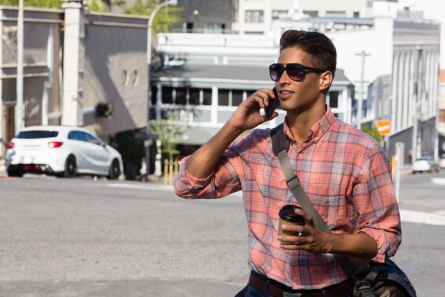 Young man in sunglasses talking on mobile phone while walking on city street, holding coffee cup. Ideal for use in lifestyle blogs, urban living articles, communication technology advertisements, and modern professional lifestyle promotions.