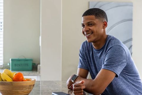 African american man leaning on kitchen countertop smiling and checking smartphone