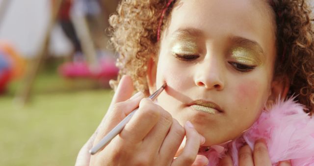 Child with curly hair is getting face painted with gold makeup during an outdoor event. Great for content related to children's activities, artistic events, parenting, outdoor festivals, creativity, and childhood experiences.
