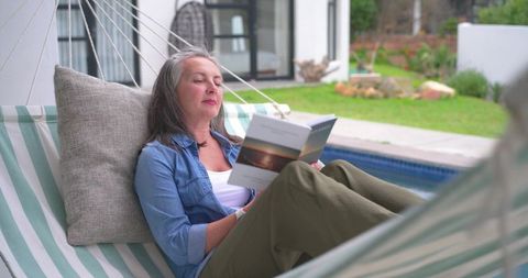 Mature woman relaxing on poolside hammock reading paperback by backyard pool and garden