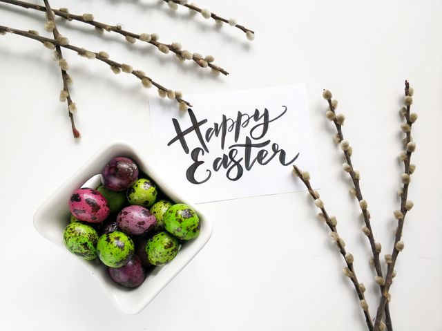 Handwritten 'Happy Easter' message next to multicolored decorative eggs in bowl and willow branches. Perfect for holiday greetings, festive decoration ideas, spring celebration themes, and Easter social media posts.
