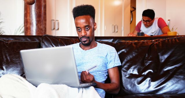Man checking information on laptop while sitting on leather sofa at home. Roommate is in background, possibly working or having a meal. Useful for illustrating home office settings, telecommuting, teamwork, daily domestic life, and modern living environments with technology.