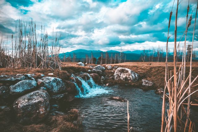 Serene mountain stream flowing amidst rocks with an expansive view of mountains. Cloudy sky creates a tranquil atmosphere. Ideal for use in nature and outdoor-themed designs, travel brochures, relaxation and meditation visuals, environmental conservation promotions.