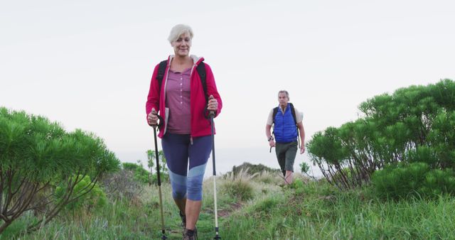 Senior hiker couple with backpacks and hiking poles while walking in the grass field. trekking, hiking, nature, activity, exploration, adventure concept.