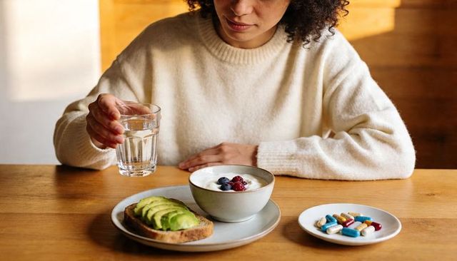 Woman reaching for daily supplements while eating avocado toast and yogurt with water