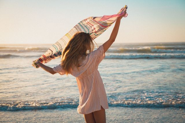 Woman enjoying a peaceful moment on the beach at sunset, holding a colorful shawl. Ideal for use in travel brochures, summer vacation advertisements, lifestyle blogs, and wellness promotions. Captures the essence of relaxation, freedom, and connection with nature.