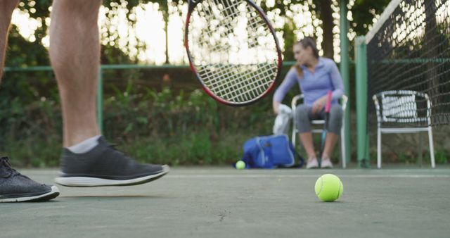 Tennis player preparing for practice on outdoor court. Close-up of tennis ball and rackets. Person seated in background organizing equipment and getting ready for game. Ideal for athletics, fitness promotions, sports equipment advertisements, and recreational activity illustrations.
