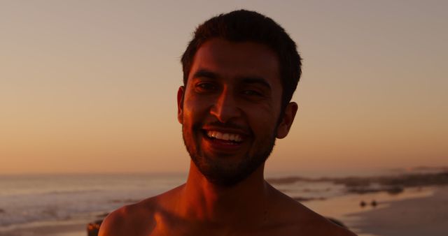 Portrait of happy biracial man with short black hair during sunset at beach. Vacation, summer and lifestyle, unaltered.