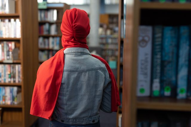Rear view of an Asian female student wearing a red hijab and jeans jacket studying in a library, standing between bookshelves.