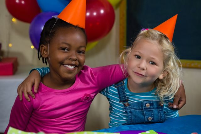 Two young girls are sitting at a table during a birthday party, wearing colorful party hats. They are smiling and have their arms around each other, showcasing their friendship and joy. The background features balloons and festive decorations, adding to the celebratory atmosphere. This image is perfect for use in advertisements, party invitations, children's event promotions, and any content related to celebrations and childhood joy.