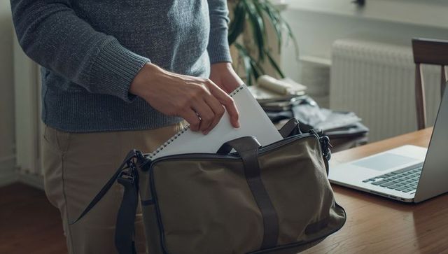 Man packing notebook into olive duffel bag on home desk with laptop, preparing for work