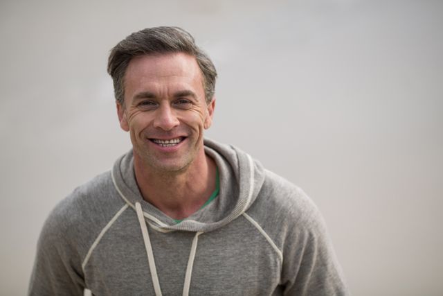 Mature man smiling while standing on the beach, wearing a casual hoodie. Ideal for use in lifestyle blogs, advertisements promoting casual wear, or articles about healthy living and happiness.