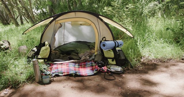 This image captures a cozy camping tent set up in a lush woodland area. The tent is surrounded by lush greenery and foliage, with various camping gear such as a sleeping bag, mat, and thermos placed in and around the tent. This image can be used to depict outdoor adventure, travel experiences, and an appreciation for nature. Ideal for websites, blogs, and promotional materials related to hiking, camping, and outdoor activities.