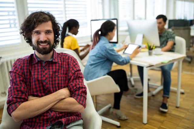 Male graphic designer sitting with arms crossed, smiling confidently in a modern office environment. Coworkers in the background working on computers and collaborating on projects. This image can be used to depict modern workspaces, team collaboration, creative industries, and professional environments.