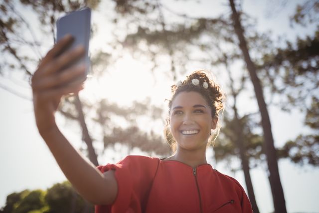 Young woman enjoying nature while taking a selfie in a forest. Ideal for use in lifestyle blogs, social media campaigns, technology advertisements, and outdoor activity promotions.