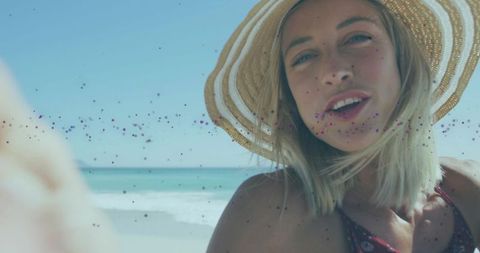 Smiling woman wearing straw hat taking selfie on sunlit beach with glitter overlay