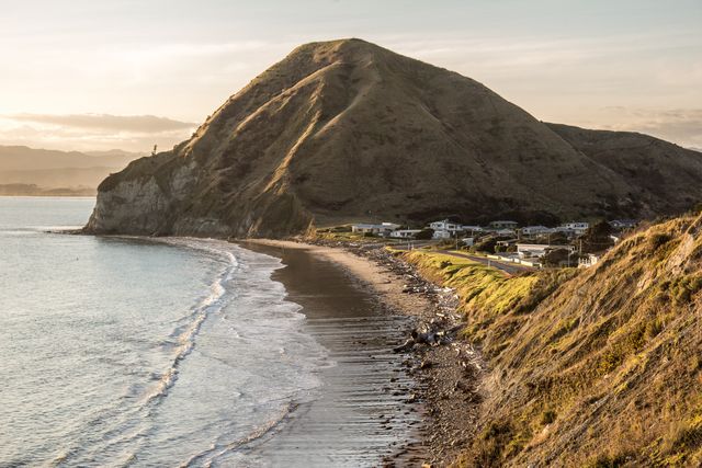 This photo captures a serene coastal scene at sunset, showcasing gently rolling hills and a quiet beach with small waves lapping at the shore. Distant homes are nestled at the base of the hills, giving a sense of isolation and tranquility. This image is ideal for use in travel promotions, nature documentaries, or lifestyle blogs focused on serene and picturesque destinations.