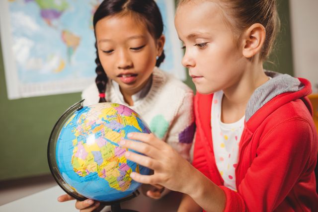Two young girls are examining a globe in a classroom, focusing on different countries. This image can be used for educational content, school promotions, geography lessons, and materials emphasizing diversity and teamwork among students.
