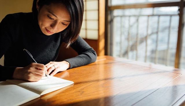 Young woman writing in notebook at sunlit wooden table leaning, journaling and concentrating