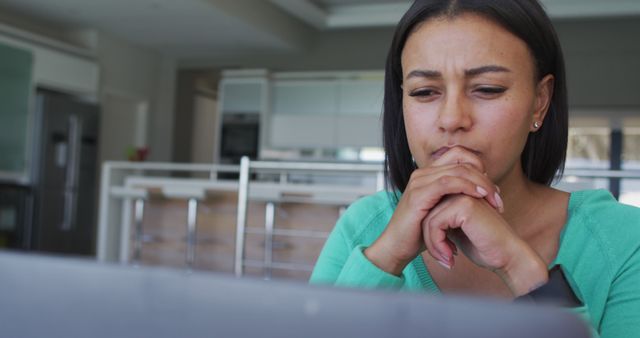 This image shows a woman deeply concentrating on a computer screen while sitting in a modern kitchen. Ideal for illustrating remote work, online study, or personal contemplation in a domestic setting. Useful for blogs, articles about home offices, focusing, or technology at home.