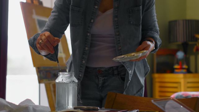 The scene captures a woman in comfortable clothing engaging in the artistic process in a home studio. She is precisely dipping a paintbrush into a jar of water while working on a canvas, suggesting concentration and creativity. Suitable for illustrating themes related to art, creativity, and at-home projects. Perfect for use in articles or promotions about hobbies, home-based artists, or painting tutorials.