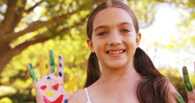 A young Caucasian girl shows off her hands painted with colorful smiley faces, with copy space. Her creative expression and joy are evident in this outdoor, playful setting.