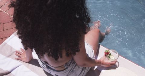 Curly-haired african american woman sitting poolside wearing denim shorts holding cocktail