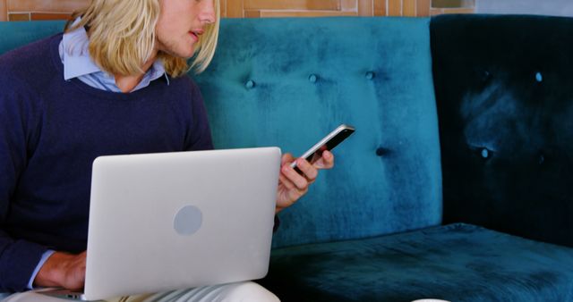 A young professional with long blonde hair working remotely, multitasking with a laptop and smartphone on a stylish blue couch. Ideal for depicting modern work environments, remote work setups, and tech-savvy professionals engaging in work and communication.