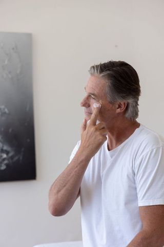 Middle-aged man applying moisturizer to face in minimalist bedroom morning routine