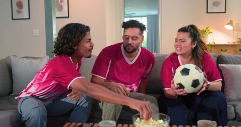 Diverse friends watching soccer on couch in red jerseys sharing popcorn holding ball