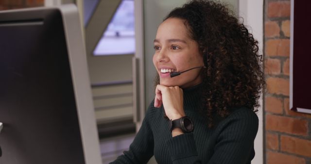 Young biracial female professional wearing headset, working at computer. She has curly brown hair, light brown skin, and is wearing dark top