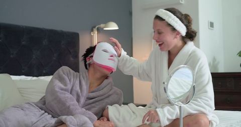 Diverse couple relaxing in robes sharing home spa night with led facial mask and mirror
