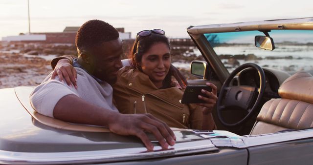A couple sitting in a convertible car, enjoying their road trip at sunset near the beach, observing something on a smartphone together. Both appear relaxed and happy. Perfect for illustrating travel adventures, romantic getaways, technology use, and carefree moments during vacations.