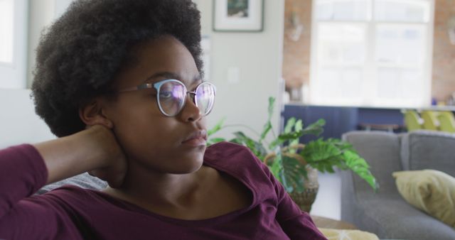 Pensive young woman wearing glasses sitting on a sofa at home. Perfect for themes about relaxation, contemplation, mental health, casual living, and work from home scenarios.