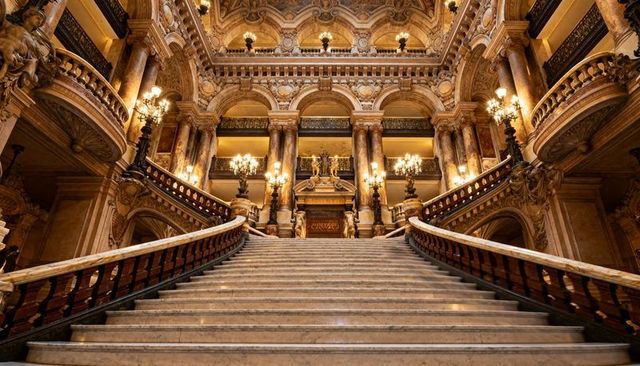 Grand neoclassical marble staircase rising through opulent theater foyer with chandeliers