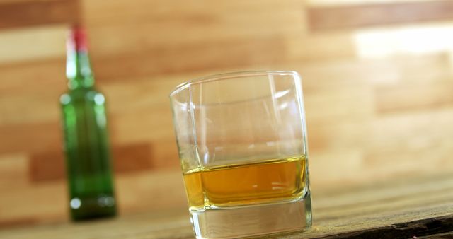 Glass of whiskey with amber hue sits on rustic wooden surface, wooden background adding warmth. Interest from focus on glass and out-of-focus bottle in the background adds depth to beverage and spirits science. Use for blogs on whiskey, advertising, or bar initiatives.