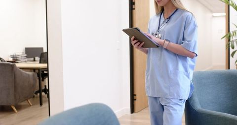 Female nurse reviewing tablet in clinic corridor wearing light blue scrubs and stethoscope
