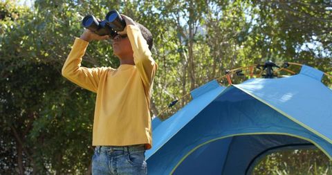 African american child exploring with binoculars beside blue tent in sunlit woodland campsite